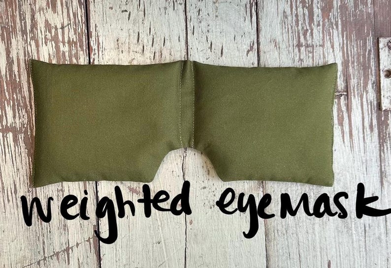 May include: An olive green weighted eye mask. The mask is rectangular with a curved cutout for the nose. The words "weighted eye mask" are written in black script below the mask. The mask is made of a soft fabric.
