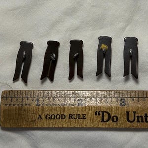 May include: Five dark brown novelty buttons shaped like trousers. Each button has a metal shank and is approximately 1 inch in length. A wooden ruler with the text "A Good Rule" and "Do Unto" is in the foreground.
