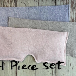 May include: A set of four fabric eye pillows in shades of pink, gray, and blue. The pink pillow has a curved cutout. The text "4 Piece Set" is written in black script below the pillows. The pillows are made of a textured fabric.