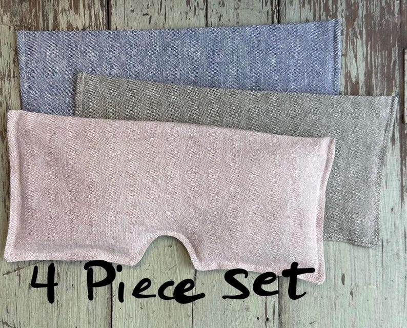 May include: A set of four fabric eye pillows in shades of pink, gray, and blue. The pink pillow has a curved cutout. The text "4 Piece Set" is written in black script below the pillows. The pillows are made of a textured fabric.