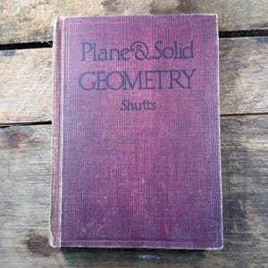 May include: A hardcover book titled Plane & Solid Geometry by Shutts with a dark maroon, textured fabric cover. The title and author name are printed in black ink on the front cover, which features a simple rectangular border design.