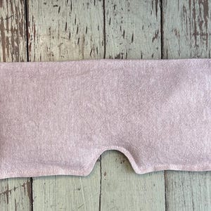 May include: A light pink eye pillow made of fabric, with a curved cutout for the nose. The rectangular pillow is filled and has a stitched edge. The background is a weathered, light green and brown wooden surface.