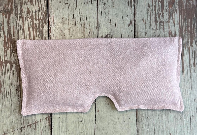 May include: A light pink eye pillow made of fabric, with a curved cutout for the nose. The rectangular pillow is filled and has a stitched edge. The background is a weathered, light green and brown wooden surface.