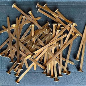 May include: A pile of brass binder clips, also known as bulldog clips, with a pointed end and a round head. The clips are arranged in a random pattern.