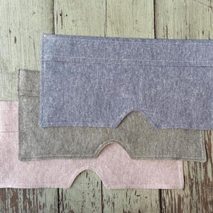 May include: Three fabric eye masks in shades of blue, gray, and pink are stacked on a weathered wooden surface. Each mask has a rectangular shape with a curved cutout for the nose, and is made of a textured material.