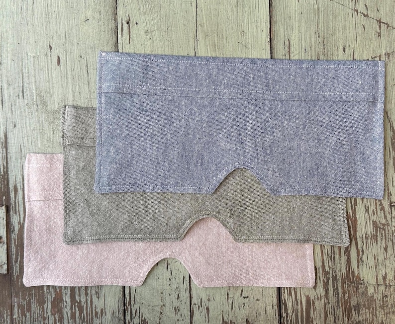 May include: Three fabric eye masks in shades of blue, gray, and pink are stacked on a weathered wooden surface. Each mask has a rectangular shape with a curved cutout for the nose, and is made of a textured material.