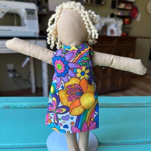 May include: A handmade doll with a white fabric body and curly blonde yarn hair. The doll is wearing a colorful dress with a floral pattern and a rainbow design.