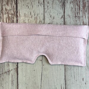 May include: A light pink eye pillow with a curved bottom edge and a raised section at the top. The pillow is made of a textured fabric and is designed to rest over the eyes.