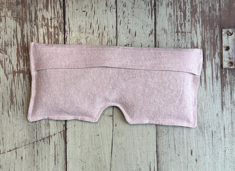 May include: A light pink eye pillow with a curved bottom edge and a raised section at the top. The pillow is made of a textured fabric and is designed to rest over the eyes.