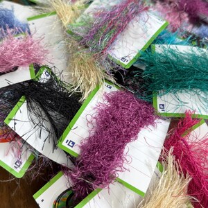 May include: A collection of multi-colored eyelash yarn skeins wrapped around white rectangular cardboard cards with green edges. The fuzzy, textured yarn comes in various vibrant shades including pink, black, purple, teal, and metallic gold.