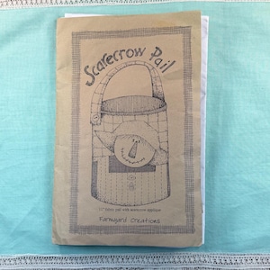 May include: A tan paper pattern envelope featuring a black line drawing of a pail with a scarecrow face applique. The text reads Scarecrow Pail, 11" fabric pail with scarecrow applique, and Farmyard Creations. The design is enclosed in a border.