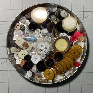 May include: A metal tin filled with a variety of buttons in different colors, sizes, and materials. The buttons include white, black, brown, gold, and clear plastic buttons.
