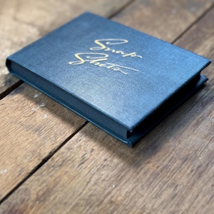 May include: A rectangular navy blue photo album with a textured fabric cover. The word Snap Shots is embossed in gold cursive script on the front cover. The album is positioned at an angle, showcasing its hardbound construction and clean edges.