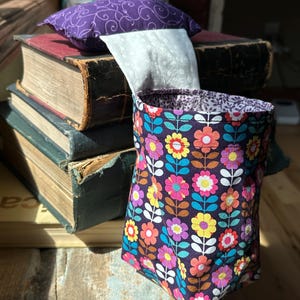 May include: A small, floral patterned fabric pouch with a purple and white fabric lining. The pouch is sitting on top of a stack of old books.