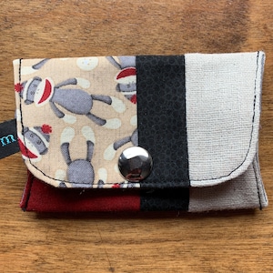 May include: A small, red, black, and white fabric wallet with a button closure. The front of the wallet features a pattern of monkeys wearing Santa hats.