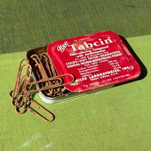May include: A vintage red metal Tabcin sliding tin container filled with several rusted metal paper clips. The lid features white text detailing the product as a pain-relieving compound for colds, hay fever, headaches, and dental work by Miles Laboratories.