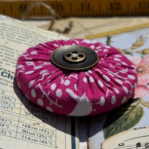 May include: A handmade, round fabric button in a vibrant pink hue, adorned with a white floral pattern. The button features a dark, antique-style metal button in the center, adding a vintage touch. This decorative button is perfect for sewing and craft projects.