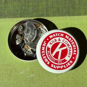 May include: A round metal tin containing assorted silver and gold-toned watch movement parts. The lid is white with a red circular logo featuring the text KILB & COMPANY WATCH MATERIAL JEWELERS SUPPLIES MILWAUKEE WIS and a large letter K.