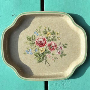 May include: A vintage serving tray with a floral design. The tray has a beige background with a green border and features a bouquet of pink and yellow roses with green leaves. The tray has a scalloped edge.