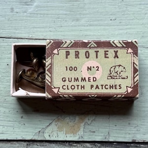 May include: A vintage box of "PROTEX" gummed cloth patches, with a bear illustration. The box is brown and green, with the drawer partially open, revealing small metal objects. The box is on a distressed, light green surface.