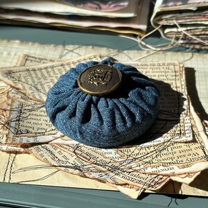 May include: A small, round denim pincushion with a decorative brass button on top. The pincushion is a deep blue color and sits on a stack of aged paper with printed text.