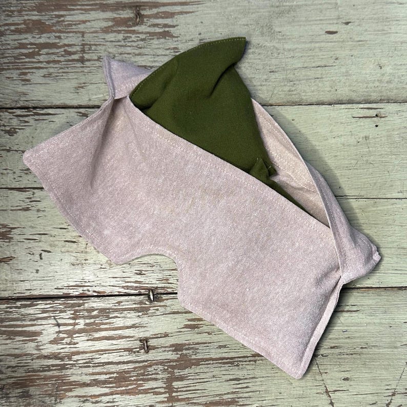 May include: A light pink fabric pouch with a green pointed hat inside. The pouch has a unique shape with a curved opening. The hat is a dark green color and is made of a soft material. The background is a light green wooden surface.