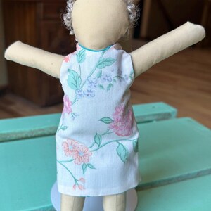 May include: A handmade doll with gray curly hair wearing a white dress with a floral pattern. The dress has a teal trim around the neckline.