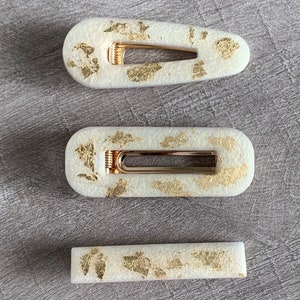 May include: Three white hair clips with gold flecks. The clips are in different sizes and shapes.