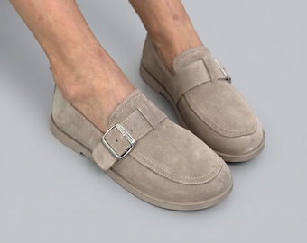 May include: A pair of light beige suede loafers featuring a single strap across the vamp with a silver-toned metal buckle closure. The shoes have a rounded toe, visible tonal stitching along the edges, and a flat, matching beige rubber sole.