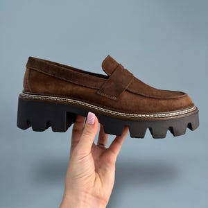 May include: Brown suede platform loafers with a chunky black sole. The shoes have a classic loafer design with a strap across the top. The sole is approximately 2 inches high. The shoes are held by a hand.
