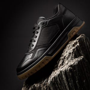 Könnte beinhalten: Ein schwarzer Sneaker mit brauner Sohle wird auf einem strukturierten, grauen Felsen vor dunklem Hintergrund gezeigt. Der Schuh hat eine Kombination aus Leder und Stoff, mit Schnürdesign und gepolstertem Kragen. Die Sohle hat ein geometrisches Muster.