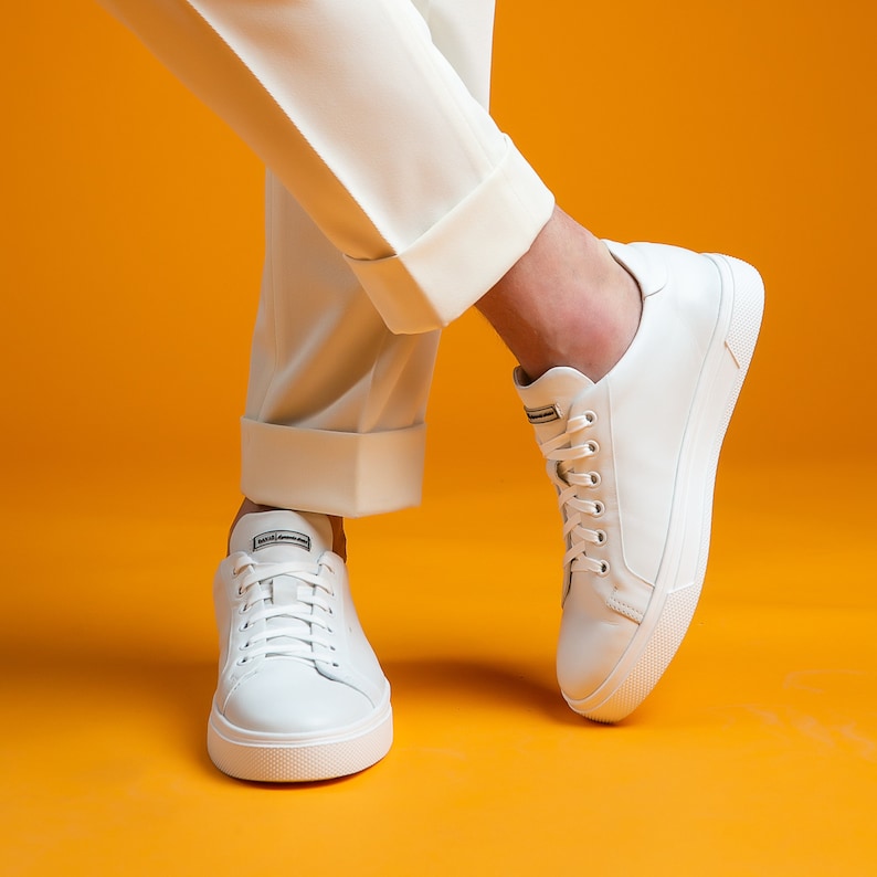 May include: White leather sneakers with white laces and a white sole. The sneakers are paired with white pants, creating a monochromatic look against a bright orange background. The sneakers have a minimalist design.