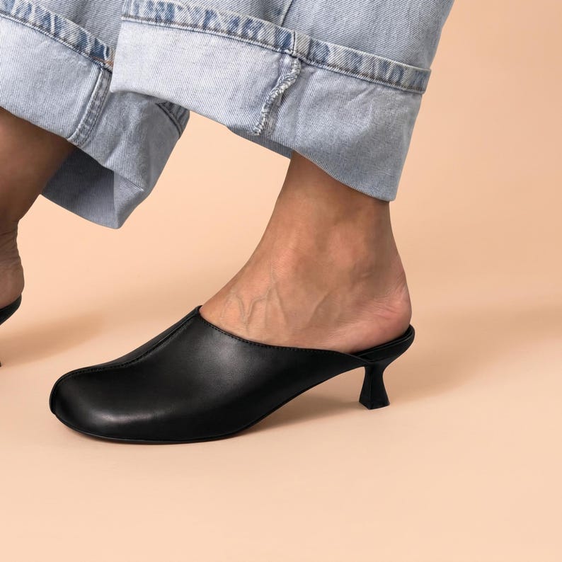 May include: A black mule shoe with a smooth finish, a rounded toe, and a central seam running down the front. The footwear features a low, flared kitten heel and an open back design, providing a slip-on style that is worn with light blue denim.