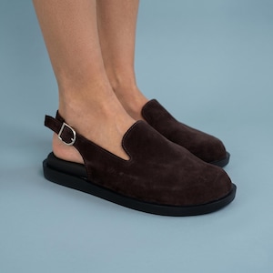 May include: A pair of dark brown suede slingback shoes featuring a rounded toe, a thick black rubber sole, and a silver-toned metal buckle on the adjustable heel strap. The footwear has a closed front design with a low-cut opening at the sides.