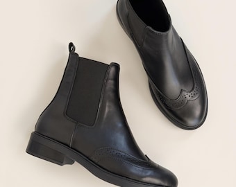 May include: A pair of black leather Chelsea boots featuring elastic side panels, a pull tab at the heel, and decorative brogue-style perforated detailing across the rounded toe cap. The boots have a low block heel and a smooth finish.
