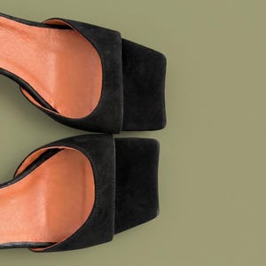 May include: Overhead shot of two black suede sandals with square toes and open sides. The interior of the sandals is a light orange color. The sandals are set against a muted green background.