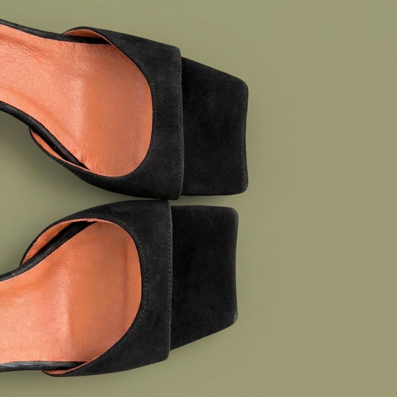 May include: Overhead shot of two black suede sandals with square toes and open sides. The interior of the sandals is a light orange color. The sandals are set against a muted green background.