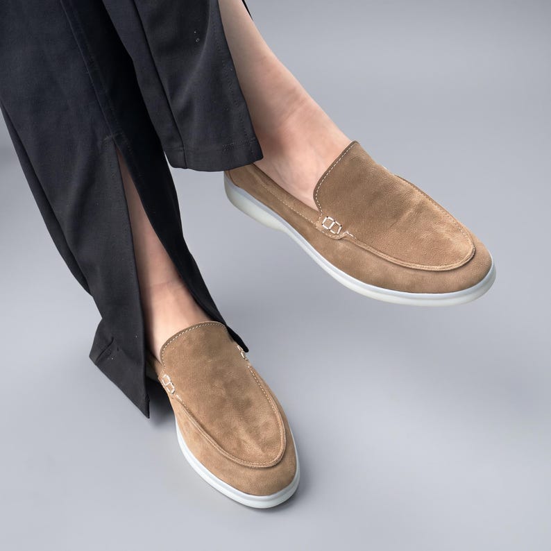 May include: A pair of tan suede loafers featuring a moccasin-style stitched toe seam, a low-profile silhouette, and a clean white rubber sole. The shoes are worn with black trousers that have a vertical slit detail at the bottom hem.