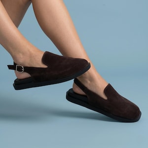 May include: A pair of dark brown suede slingback shoes featuring a closed rounded toe, a flat black sole, and an adjustable heel strap with a small silver-toned metal buckle. The shoes have a simple slip-on design with a soft textured finish.