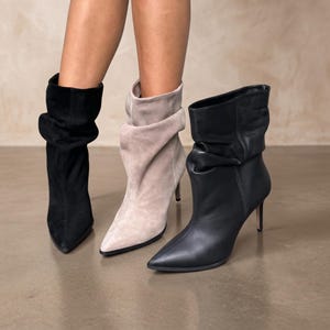 Op de afbeelding: Drie enkellaarsjes met spitse neuzen en stilettohakken. Eén laars is van zwart suède, één van lichtbeige suède en één van glad zwart leer. Elke laars heeft een soepel, geplooid schachtontwerp dat tot halverwege de kuit reikt.