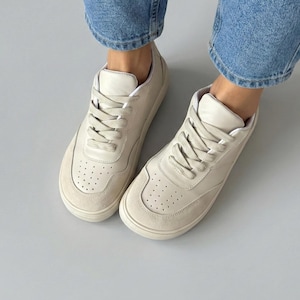 May include: Cream-colored sneakers with a suede toe cap and a perforated design. The shoes have white laces and a white sole. The sneakers are worn with blue jeans.