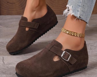 May include: A pair of dark brown suede clogs featuring a rounded toe, a low-profile silhouette, and a single adjustable strap across the instep secured with a silver-toned metal buckle. The shoes have a flat black sole and a smooth, matte finish.