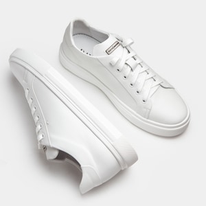 May include: Two white leather sneakers with white laces and soles. The sneakers have a low-profile design and a small label that reads "DAVIS". The shoes are set against a white background.