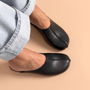 May include: A pair of black leather slip-on mules featuring a rounded toe design and a prominent center seam running from the opening to the front. The footwear has a smooth, matte finish and a low-profile silhouette that covers the top of the foot.