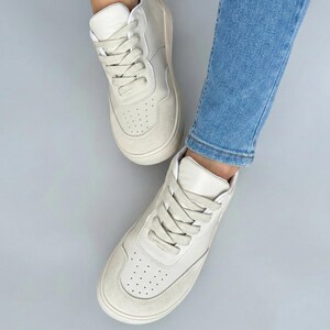 May include: Off-white high-top sneakers with matching laces. The sneakers have a suede toe and a perforated design on the front. The shoes are worn with blue jeans.