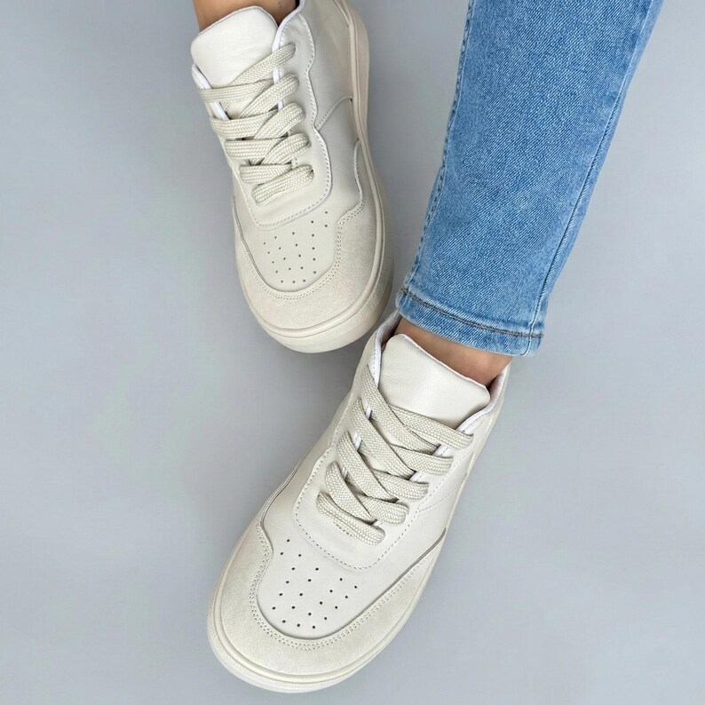 May include: Off-white high-top sneakers with matching laces. The sneakers have a suede toe and a perforated design on the front. The shoes are worn with blue jeans.