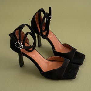 May include: A pair of black suede heeled sandals with square toes and ankle straps. The sandals have a high, block heel and a silver buckle closure. The interior of the shoe is a light brown color. The shoes are set against a muted green background.
