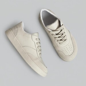 May include: A pair of low-top sneakers in a solid off-white color. The shoes feature a smooth upper with a textured toe cap, matching thick laces, and a perforated detail on the front. The soles are flat and textured with a rubberized finish.