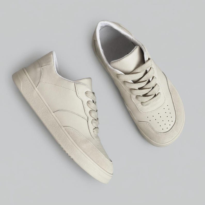 May include: A pair of low-top sneakers in a solid off-white color. The shoes feature a smooth upper with a textured toe cap, matching thick laces, and a perforated detail on the front. The soles are flat and textured with a rubberized finish.