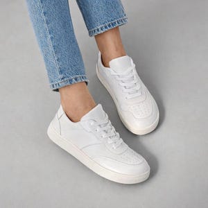 May include: A pair of clean white sneakers featuring a low-top silhouette, matching white laces, and a perforated toe box design. The shoes are constructed with a smooth finish and a thick, solid white rubber sole for a classic look.